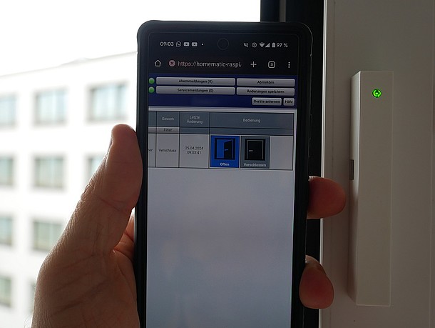 rechts weißer, länglicher Fensterkontakt mit grüner LED am Fensterrahmen, mittig ein Smartphone mit App-Anzeige für geöffnetes Fenster, links im Hintergrund ein Gebäude mit vielen Fenstern